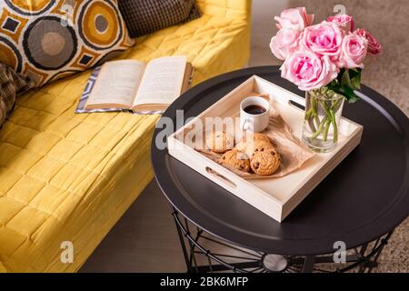 Home still life with bunch of pink roses, homemade snack and drink on small table and open book and cushions on yellow couch Stock Photo