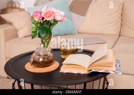 Small table with rose bunch in glass of water, green tea in teapot and cup and open book of stories against couch with pillows and tablet Stock Photo