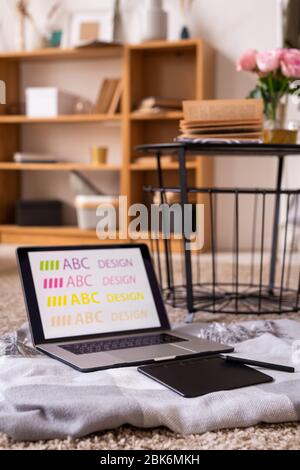 Pad with stylus and laptop of designer on the floor of living-room with small table with open book and pink roses standing near by Stock Photo