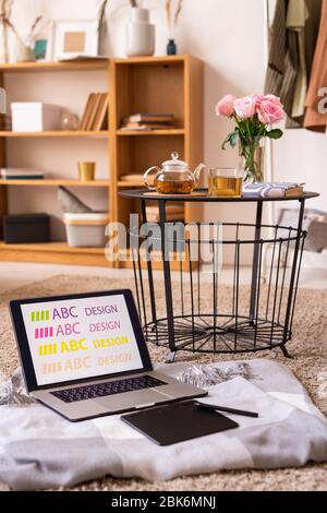Laptop and pad with stylus on plaid by small table with herbal tea and pink roses standing on the floor with bookshelf on background Stock Photo