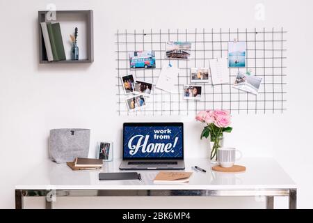 White desk with laptop, pad with stylus, notepads, sketchbook, bunch of pink roses, mug with drink, shelf with books and photos above on wall Stock Photo