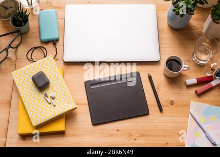 Notebooks with cordless earphones, folded laptop, pad with stylus, cup of coffee, glass of water, lip gloss, pen and domestic plants on table Stock Photo