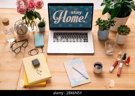 Stay home message on display of laptop on workplace of modern female designer or student with books, flowers, drinks and cosmetics Stock Photo