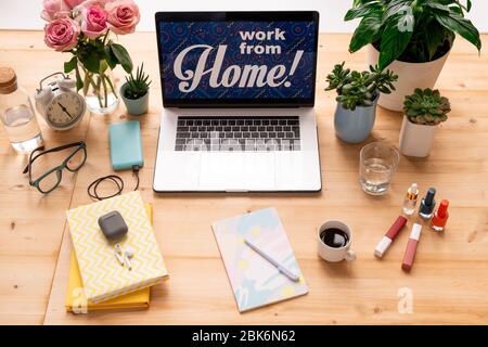 Laptop with notice work from home on display surrounded by flowers, books, copybook with pen, makeup items, drinks and alarm clock Stock Photo