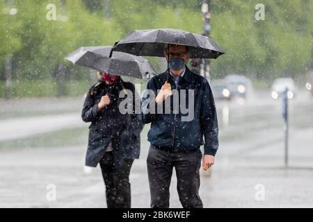 Berlin, Germany. 02nd May, 2020. A man and a woman walk in heavy rain with mouth and nose protectors and umbrellas over the 18th March square. Credit: Christoph Soeder/dpa/Alamy Live News Stock Photo