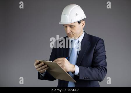 Chief engineer in hard hat makes notes on clipboard isolated on grey background Stock Photo