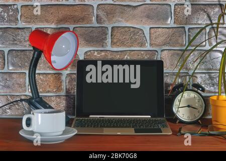 Close up of workplace with laptop, glasses, coffee cup, table lamp and alarm clock. Stock Photo