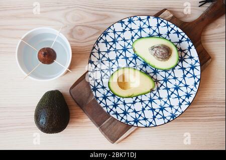Top view of avocado fruit on ceramic vintage dish and seed inside pot for growing a plant. Avocado seed in water for reproduction of seedlings. Health Stock Photo