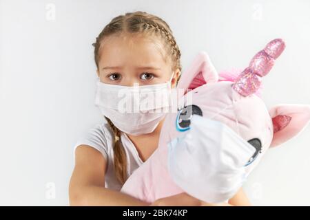 Child in medical mask hugs a soft pink masked unicorn on a white background. Stock Photo