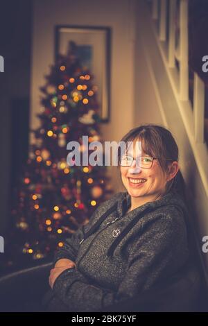 Close up of cheerful lady sitting in chair, relaxing in front of decorated Christmas tree looking pleased after cooking & eating the Christmas dinner. Stock Photo