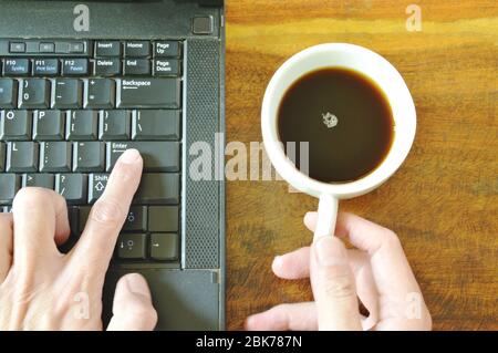finger pushing enter button on computer notebook while holding coffee cup Stock Photo