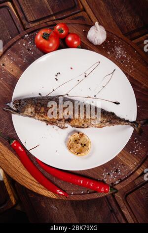 Appetizing mackerel fish baked on fire with spices, served on a plate and decorated with vegetables on wooden background Stock Photo