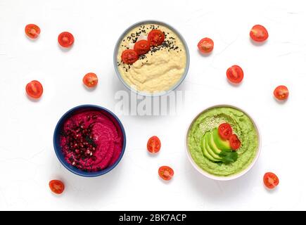 hummus consisting of beets, avocados and green peas, chickpeas in bowls. on a white background decorated with tomatoes. copy space. flat lay Stock Photo