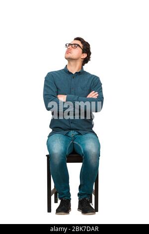 Full length portrait of puzzled businessman seated on a chair, wearing eyeglasses and keeps arms crossed, looking up perplexed, isolated on white back