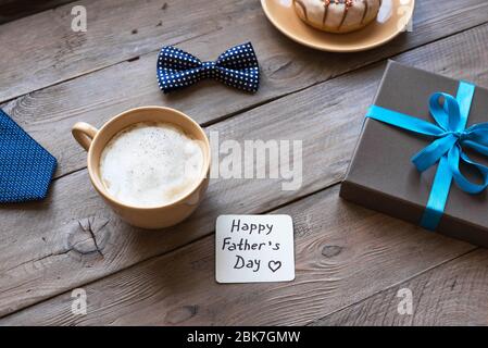 Happy Fathers Day composition on wooden table. Morning coffee, donut, gift box with greeting card, Father's Day celebration concept. Stock Photo