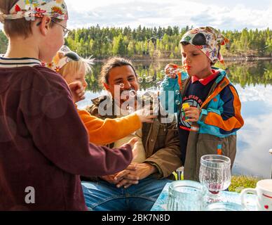 Two boys wearing patterned headscarves are blowing soap bubbles at a picnic table by the lakeside, whilst a man in a brown fleece jacket sits between them, laughing. There are glasses and a cup on the table. Armaantie, Ruuhijärvi, Nastolan suuralue, Lahti, Lahti sub-region, Päijät-Häme, Mainland Finland, 15580, Finland Stock Photo