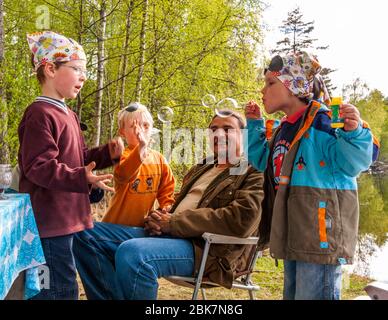At a picnic table by the lakeside, a man in a brown fleece jacket is sitting amongst several children, whilst a boy wearing a patterned headscarf blows a soap bubble from a tube. In the background, the lake lies in front of a birch forest. Armaantie, Ruuhijärvi, Nastolan suuralue, Lahti, Lahti sub-region, Päijät-Häme, Mainland Finland, 15580, Finland Stock Photo