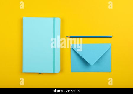 Neatly organized stationery flat lay of pastel blue notebook, envelope and pencil over yellow paper background, elevated top view, directly above Stock Photo