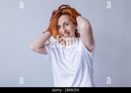 Horizontal portrait of pleasant-looking Caucasian female with long redhair,  wearing white casual T-shirt,  looking happily in camera Stock Photo