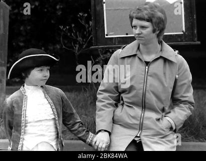 The annual Knutsford Royal May Day procession in 1976 in Knutsford, Cheshire, England, United Kingdom. It traditionally includes a fancy-dress pageant of children in historical or legendary costumes with  horse-drawn carriages. A woman holds the hand of a young boy dressed in a historical costume, waiting for the procession to begin. Stock Photo