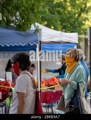 Davis, California, USA. May 2, 2020. Masked custommers wait in line at the  farmers, which has remained open despite the lockdown. The tents are  more spread out and only the vendors can touch the produce. Money and produce are handled by different people. A hand washing stations is offered and almost everyone is wearing masks. Stock Photo
