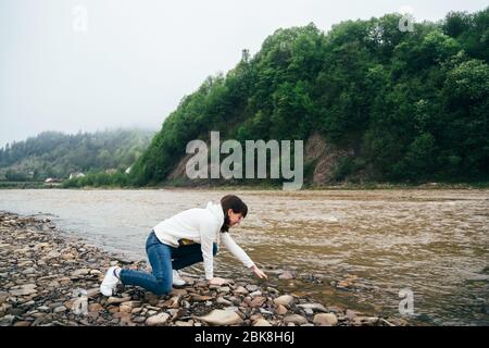 Tourist girl in jeans and white sweatshirt touching the yellow water on the bank of the dirty mountain river surrounded by forest Stock Photo