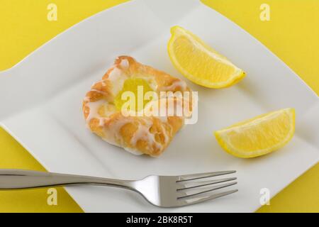 Mini lemon danish breakfast pastry with fresh lemon wedges on white plate with fork on yellow background Stock Photo
