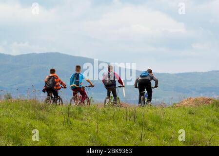 A group of four children riding a bicycle among nature against a mountain background. Тhey goes fishing through a green meadow. Stock Photo
