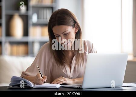 Smiling woman writing in notebook, using laptop at home Stock Photo
