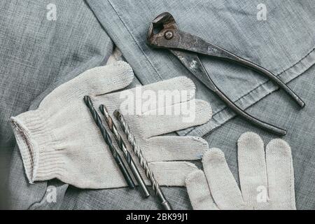 A handful of metal drill bits and rusty pliers lies on Safety knitted Work Gloves for industrial use against the backdrop of an old denim fabric Stock Photo