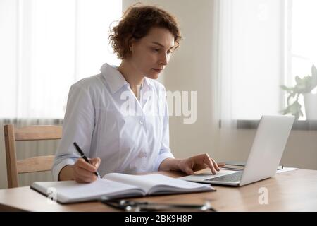 Young woman doctor working on laptop taking notes at workplace Stock Photo
