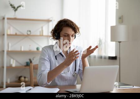 Female general practitioner wears headset video conferencing on laptop Stock Photo