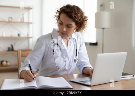 Serious female doctor working using laptop and writing notes Stock Photo