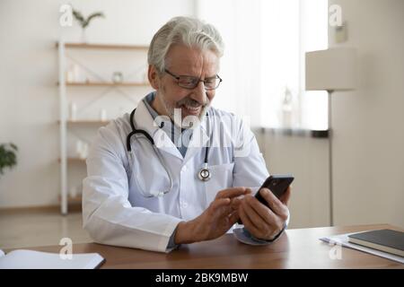 Happy old doctor holding using smart phone in medical office Stock Photo
