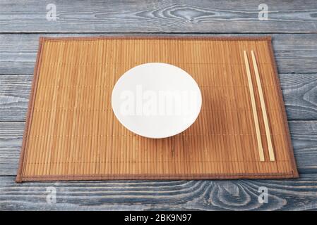 A white empty bowl and two wooden chopsticks lie on a bamboo mat that sits on a wooden table made of textured boards. Stock Photo