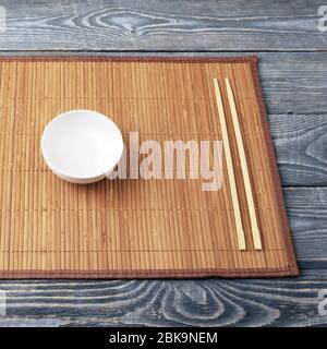A white empty bowl and two wooden chopsticks lie on a bamboo mat that sits on a wooden table made of textured boards. Stock Photo