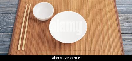 Two white empty bowl and two wooden chopsticks lie on a bamboo mat that sits on a wooden table made of textured boards. Stock Photo