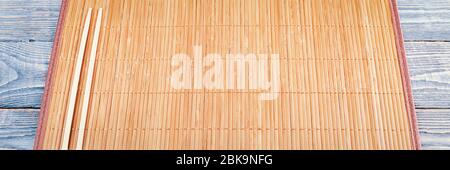 Two wooden chopsticks lie on the edge of a bamboo Mat that sits on a wooden table made of textured boards. Stock Photo