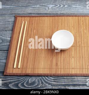 A white empty bowl and two wooden chopsticks lie on a bamboo mat that sits on a wooden table made of textured boards. Stock Photo