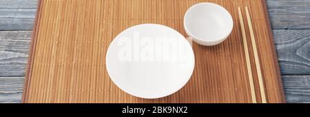 Two white empty bowl and two wooden chopsticks lie on a bamboo mat that sits on a wooden table made of textured boards. Stock Photo