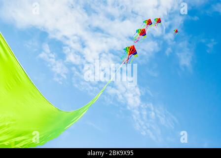 Rainbow colored kites flying high on the end of a vibrant green ribbon against a Summer blue sky and light clouds Stock Photo