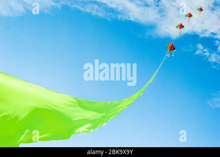 Rainbow colored kites flying high on the end of a vibrant green ribbon against a Summer blue sky and light clouds Stock Photo