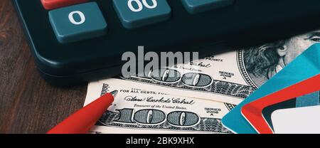 A black calculator, hundred-dollar bills, a bright red ballpoint pen, and multicolored credit cards fanned out on a brown wooden table. Concept of ban Stock Photo