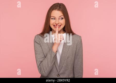 Shh, don't tell anyone! Portrait of happy young woman in business suit smiling and holding finger on lips showing silence gesture, shushing asking to Stock Photo