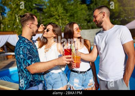 Group of friends having fun at poolside summer party clinking glasses with summer cocktails on sunny day near swimming pool. People toast drinking Stock Photo