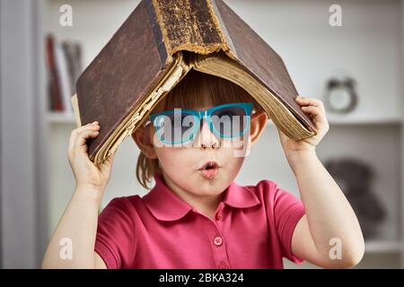 Cute little girl with glasses with an old antique book over her head. Intimate knowledge. Stock Photo