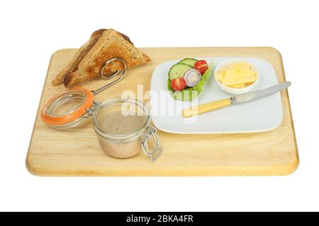 Pate in a jar with toast in a rack and salad on a plate with a knife on a wooden board isolated against white Stock Photo