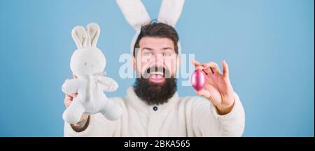 Celebrating the new life. Hipster with long rabbit ears holding egg laying hare. Bearded man with bunny toy and Easter egg. Celebration of spring time holiday. Easter bunny delivering colored eggs. Stock Photo