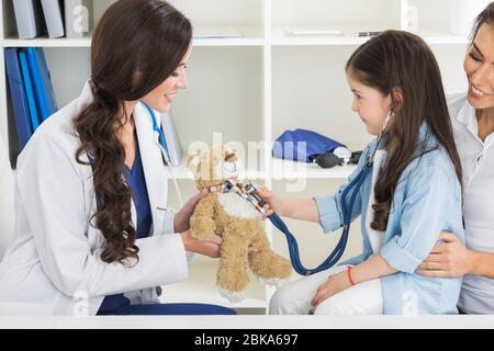 Cute small child patient pretending to be doctor, holding teddy bear toy at meeting with general practitioner. Friendly nurse showing how stethoscope Stock Photo