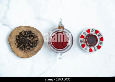 Dry black tea leaf in wooden platter and traditional Turkish tea glass with a teapot on marble background. Traditional, healthy and organic beverage. Stock Photo